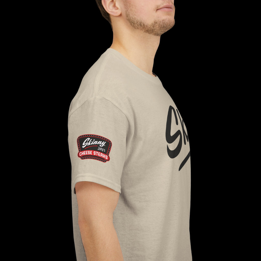 Beige t-shirt with a logo on the sleeve and text on the chest, worn by a person.