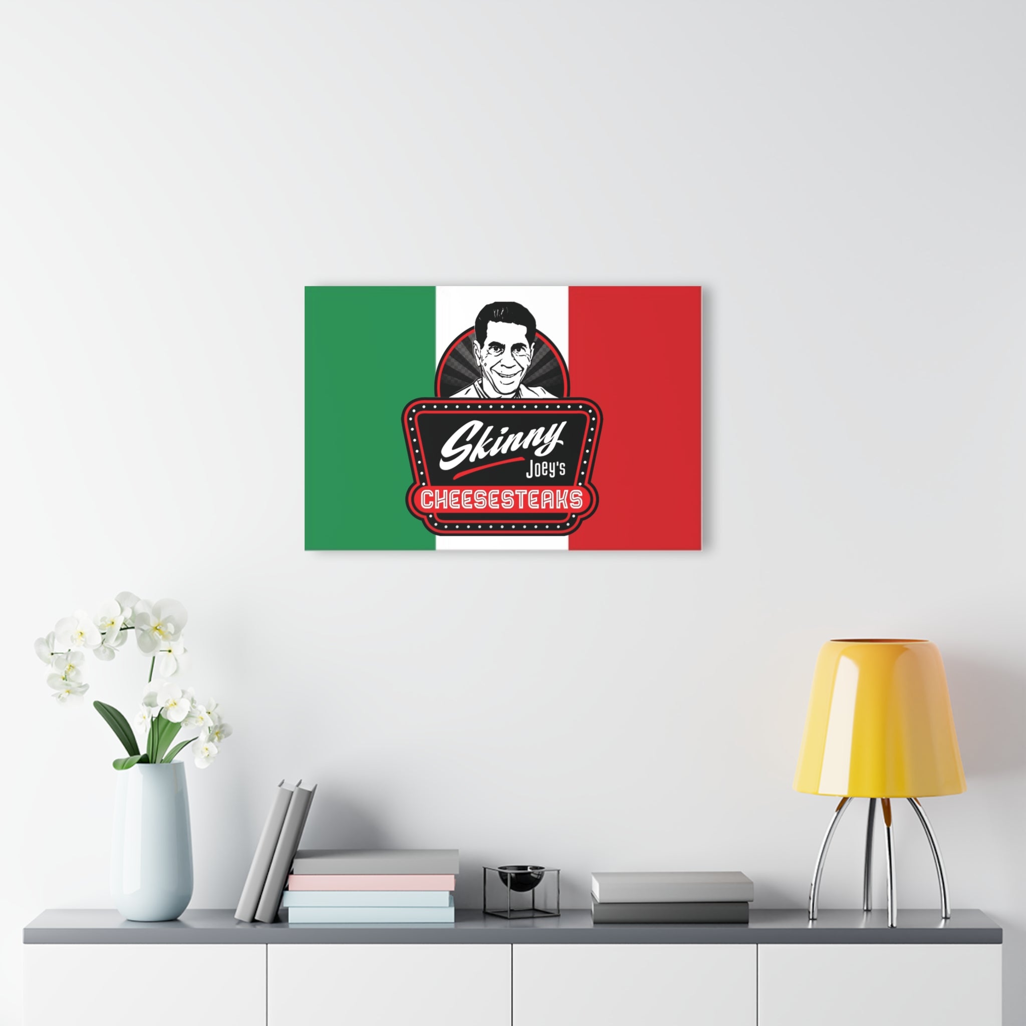 Canvas print with a stylized face and 'Skinny Joey's Cheesesteaks' text on an Italian flag background, displayed on a wall above a shelf.