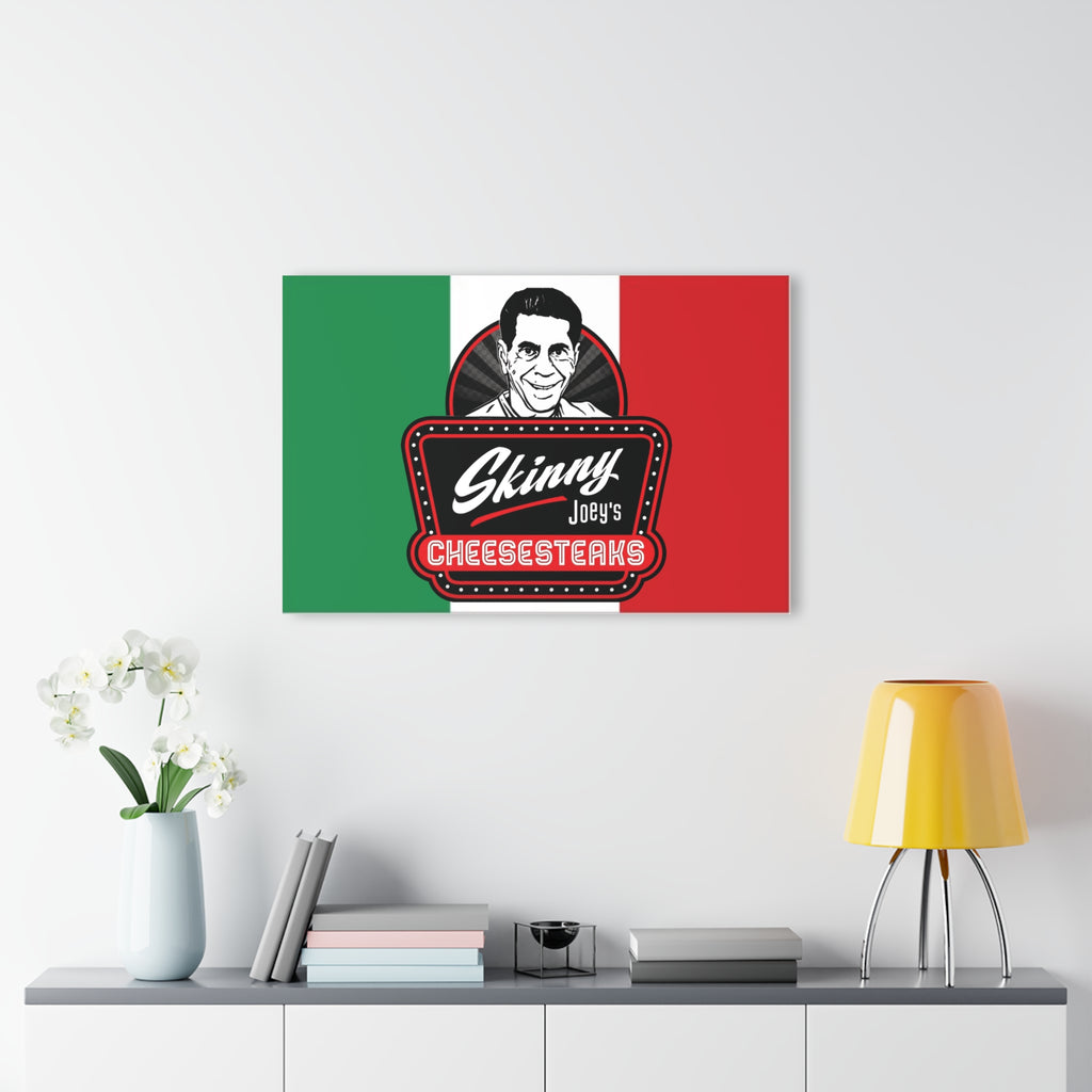 Canvas print with a stylized face and 'Skinny Joey's Cheesesteaks' logo on a green, white, and red background, displayed above a table with decor items.