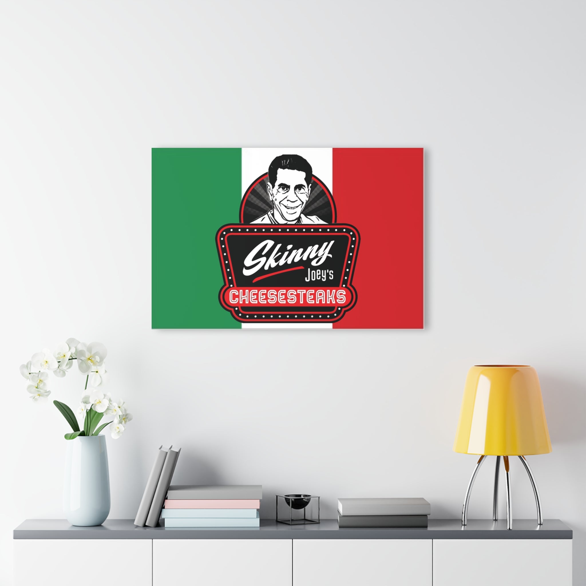 Canvas print with a stylized face and 'Skinny Joey's Cheesesteaks' logo on a green, white, and red background, displayed above a table with decor items.