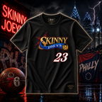 A black short-sleeve graphic T-shirt centered in the frame, featuring a large 'Skinny Joey's' logo with gold, blue, and red elements and the number 23 in white with a red outline. The shirt is shown against a dark, dramatic city street backdrop with neon signs, a basketball, and a flag.